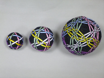 A photographic image of three balls in a line, growing in size from left to right. All three balls are black and have five lines strewn onto them, where the complexity of the line's patterns also grows from left to right.
