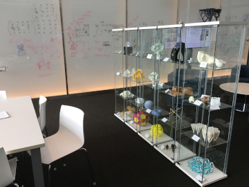 A working space at ICERM with a table and chairs on the left, a large whiteboard in the background with some mathematical notation on it, and a glass case on the right, exhibiting various illustration examples of mathematical objects.